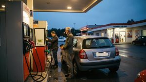 Un homme fait le plein d’une voiture compacte dans une station-service française au crépuscule, avec des pompes éclairées et un panneau d’affichage vierge.